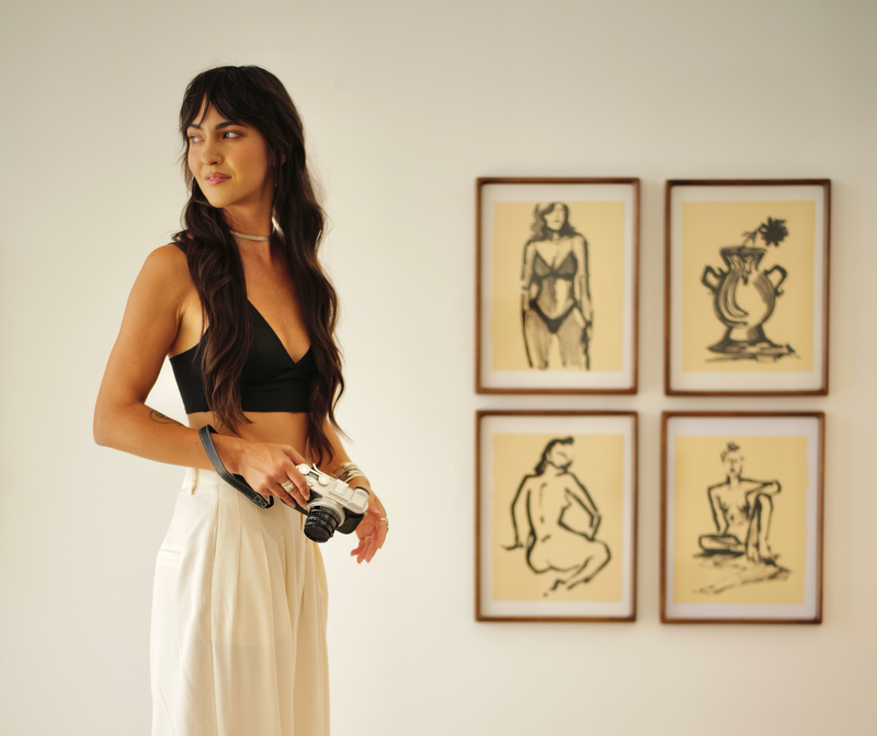 Woman standing on a stool holding a camera in front of framed artwork on a wall.